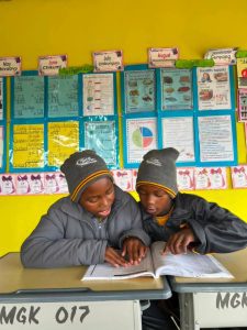 Primary school students sitting together at a desk, focused on reading their books.